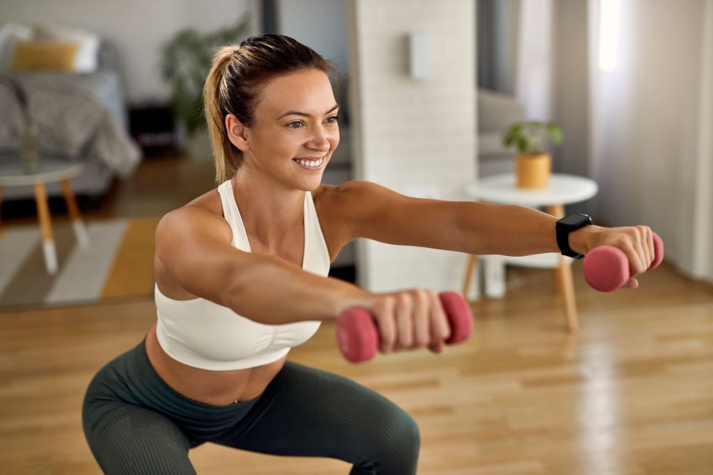 Exercícios Terapêuticos para Fibromialgia: Conheça 6 benefícios para melhorar sua qualidade de vida 3 young happy sportswoman doing squats while exercising with hands weights living room 1